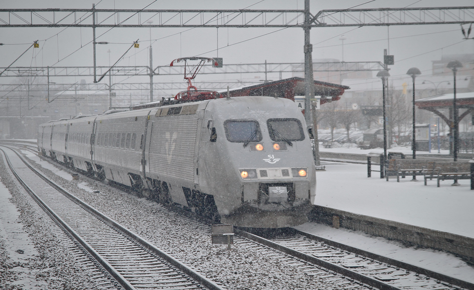 Ett snötäckt tåg på en perrmed lyset på på en perrong. Det snöar ute.