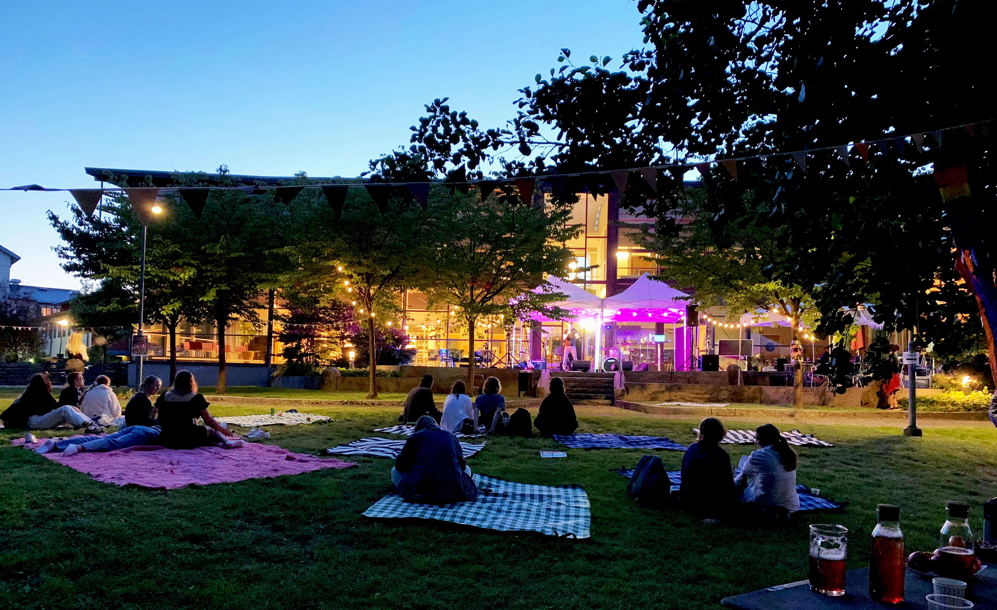 Sen kväll bakom kulturhuset i Mölnlycke med en färgsatt scen och publik på picknickfiltar bland träden.