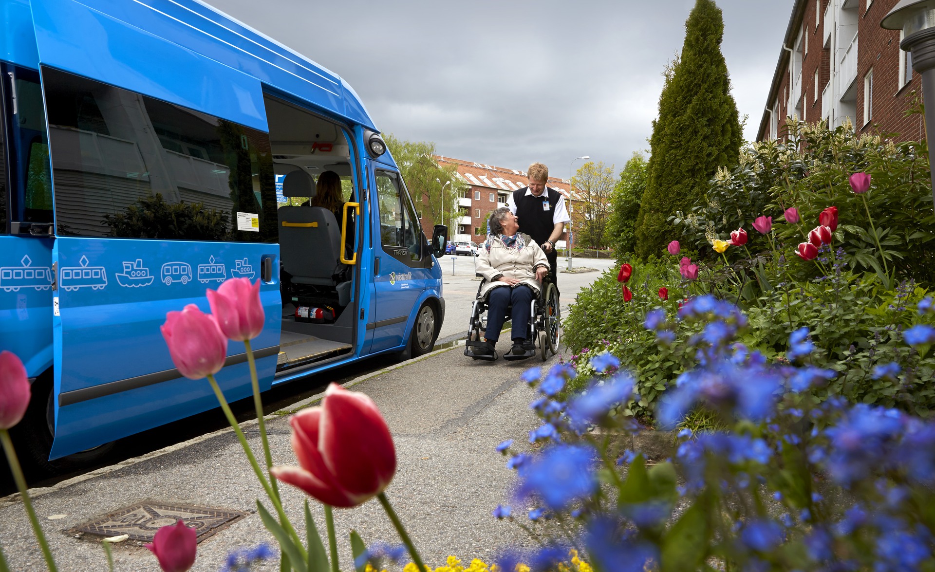 En blå buss är parkerad vid en trottoar, utanförsitter en kvinnlig resenär i rullstol, bakom hennes rygg står en busschaufför som är beredd att hjälpa kvinnan med resan.
