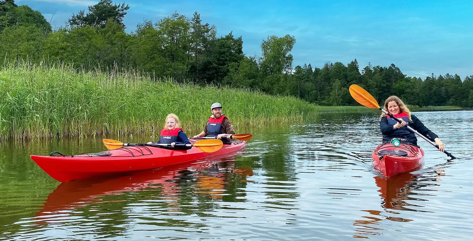 Två röda kajaker på spegelblank sjö med vass i bakgrunden. I den ena kajaken sitter ett barn och en man och i den andra en kvinna.